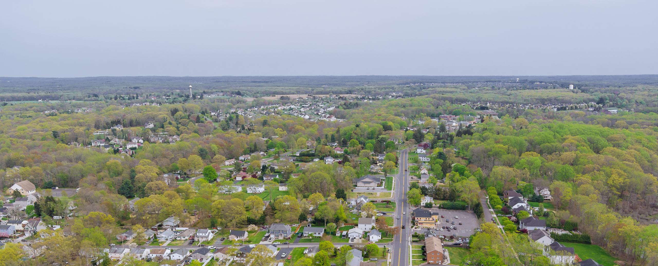 An aerial view over a residential town, with houses and tree-lined streets along a main road