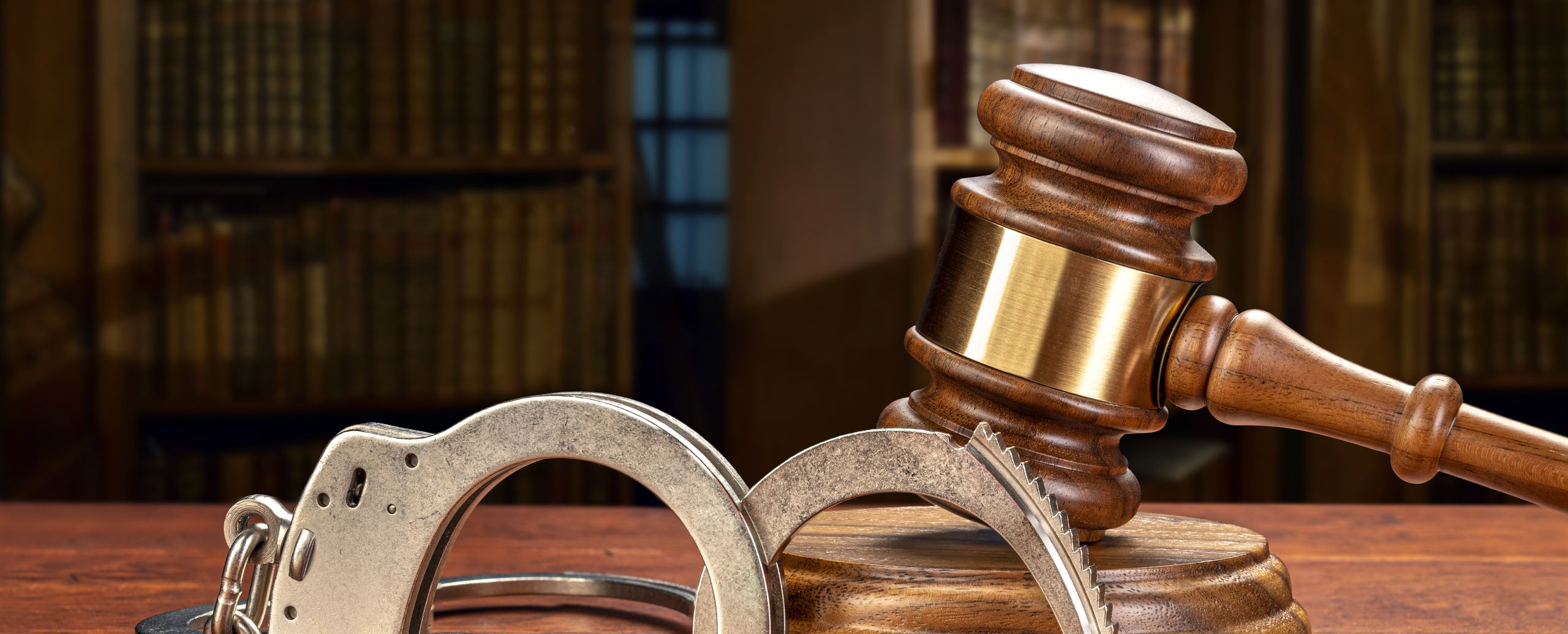 A judge&rsquo;s gavel and a pair of handcuffs on a desk in front of a law library