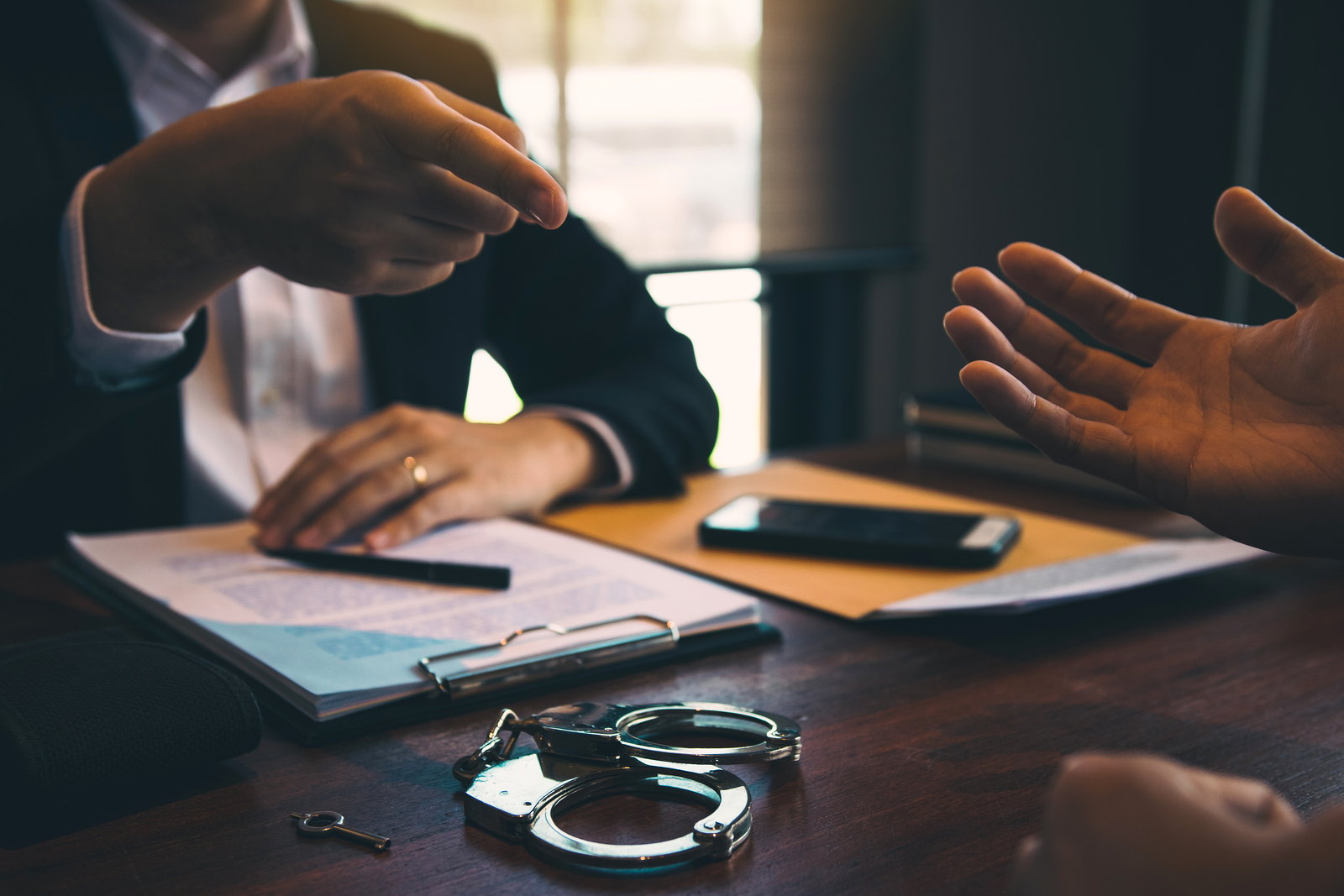 Hands across a desk in a meeting with a lawyer, handcuffs and case papers on the table